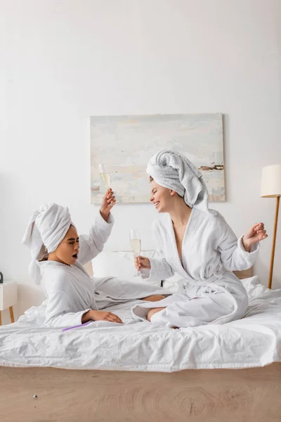 Excited african american woman toasting with champagne near happy friend in white robe and towel in bedroom - foto de stock
