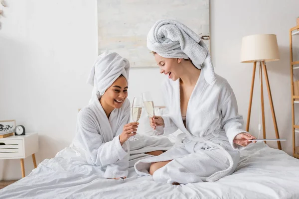 Gai interracial femmes en blanc doux peignoirs et serviettes clinquant verres à champagne sur le lit — Photo de stock