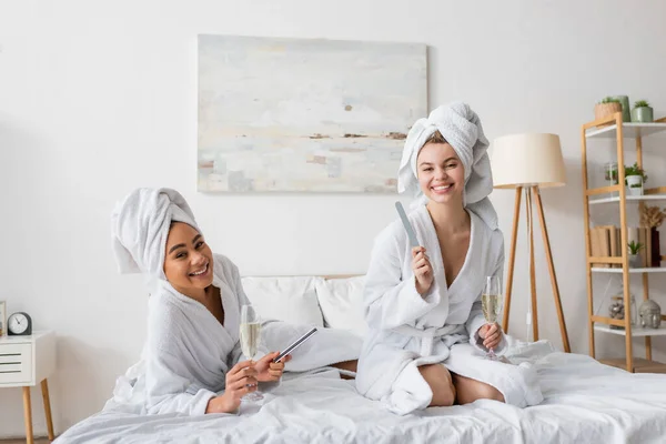 Cheerful interracial women with champagne glasses and nail files looking at camera on bed - foto de stock