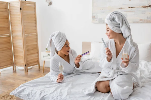 Happy multiethnic women with champagne glasses and nail files talking on bed at home — Photo de stock