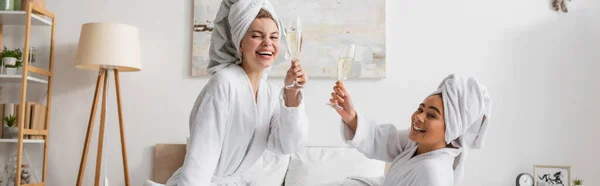 Joyful multiethnic women in white bathrobes and towels toasting with champagne glasses and looking at camera in bedroom, banner — Foto stock