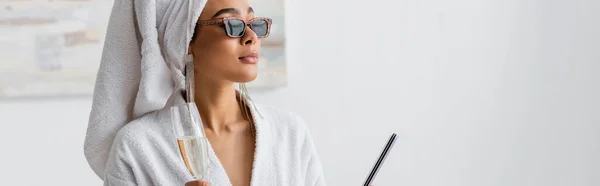 African american woman in white robe and stylish sunglasses holding champagne and nail file while looking away at home, banner — Stockfoto