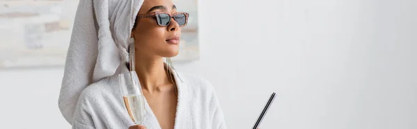 African american woman in white robe and stylish sunglasses holding champagne and nail file while looking away at home, banner — Foto stock