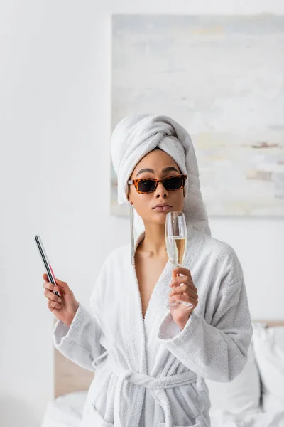 Young african american woman in white bathrobe and stylish sunglasses holding champagne glass and nail file in bedroom — Photo de stock