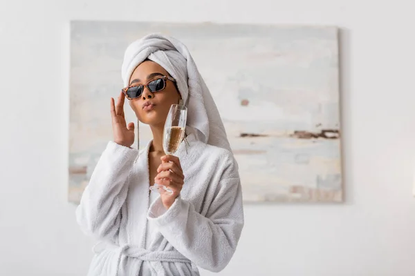 Afroamerikanerin in Frottee-Bademantel und Handtuch, die trendige Sonnenbrille anpasst, während sie Lippen schmollt und Champagner im Schlafzimmer hält — Stockfoto