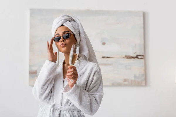 African american woman in terry bathrobe and towel adjusting trendy sunglasses while pouting lips and holding champagne in bedroom — Stock Photo