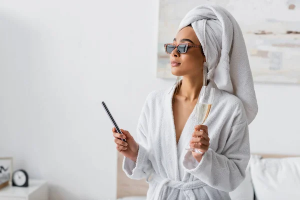 Young african american woman in soft terry bathrobe and sunglasses looking away while holding nail fail and champagne in bedroom — Stockfoto