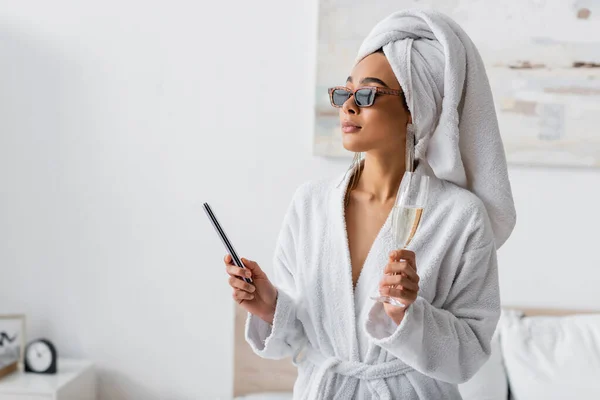 Young african american woman in soft terry bathrobe and sunglasses looking away while holding nail fail and champagne in bedroom — Foto stock