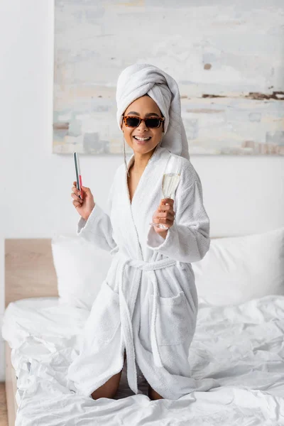 Cheerful african american woman in white bathrobe and sunglasses posing with nail file and champagne glass in bedroom — Foto stock
