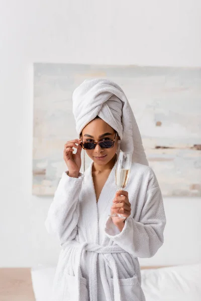 Young african american woman in white bathrobe and towel holding champagne and looking at camera over sunglasses in bedroom — Stockfoto