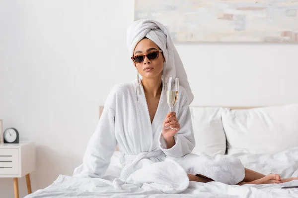 Barfüßige Afroamerikanerin in weißem weichen Handtuch und stylischer Sonnenbrille mit Champagnerglas auf dem Bett sitzend — Stockfoto