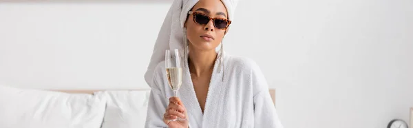 Young african american woman in white bathrobe and sunglasses with earrings holding champagne glass in bedroom, banner — Stockfoto