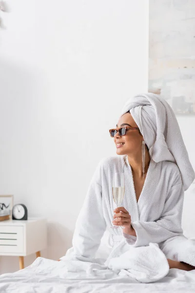 Lächelnde Afroamerikanerin im Frottee-Bademantel und trendiger Sonnenbrille, die Champagner hält und im Schlafzimmer wegschaut — Stockfoto