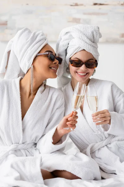 Lächelnde interrassische Frauen in weißen weichen Bademänteln und trendigen Sonnenbrillen klirren Champagnergläser im Schlafzimmer — Stockfoto