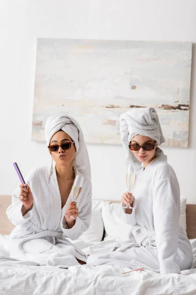 Giovani donne interrazziale in accappatoi bianchi e occhiali da sole alla moda in posa con bicchieri di champagne e lima per unghie sul letto — Foto stock