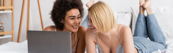 Blonde and brunette interracial women smiling near laptop in bedroom at home, banner — Stock Photo