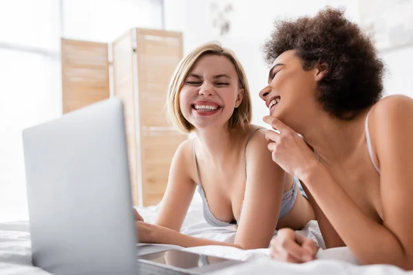 Excited multiethnic women in bras laughing near blurred laptop on bed at home — Stock Photo