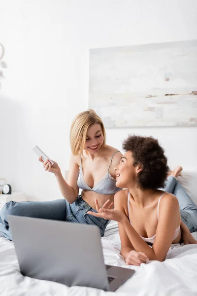 Blonde woman holding smartphone near smiling african american friend pointing at laptop on bed — Stock Photo