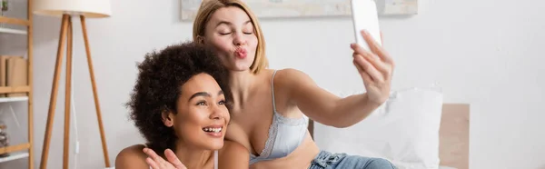 Blonde woman with closed eyes pouting lips while taking selfie on smartphone with african american friend, banner — Stock Photo