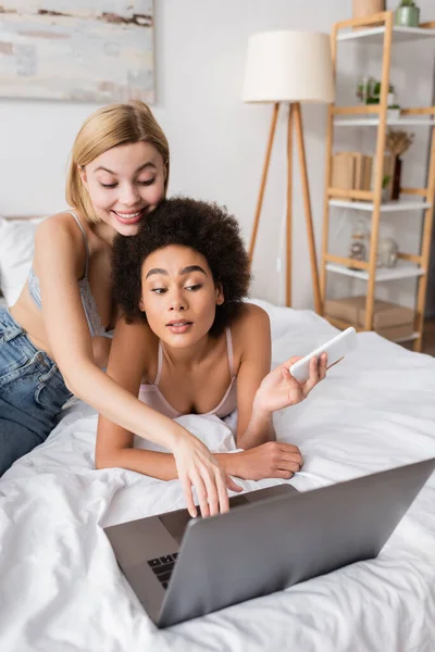 African american woman holding smartphone near smiling blonde friend using laptop on bed — Stock Photo