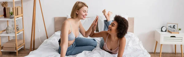 Heureux interracial femmes en soutiens-gorge et jeans donnant haute cinq sur le lit dans la chambre moderne, bannière — Photo de stock