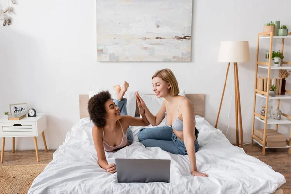 Joyful multiethnic women in lingerie and jeans giving high five near laptop in modern bedroom — Stock Photo