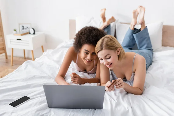Young blonde woman holding credit card near african american friend and laptop in bedroom — Stock Photo