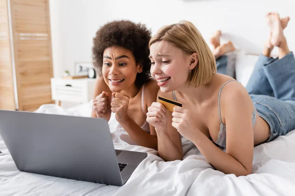 Tense multiethnic women holding clenched fists for luck near credit card and laptop in bedroom — Photo de stock