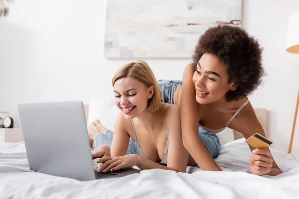 Cheerful african american woman holding credit card near blonde friend using laptop on bed — Stock Photo