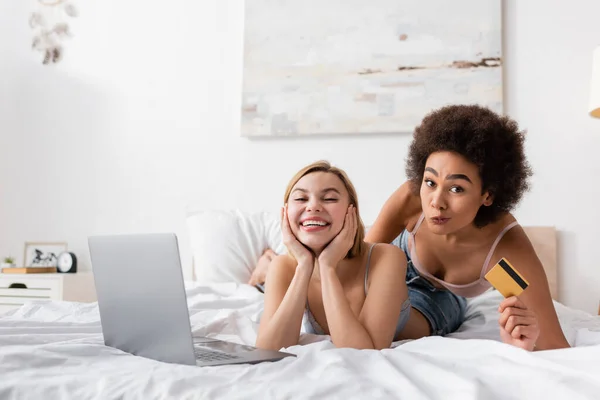 African american woman holding credit card and grimacing near cheerful blonde friend and laptop on bed — Stock Photo