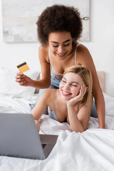 Young african american woman holding credit card near happy blonde woman looking at blurred laptop in bedroom — Fotografia de Stock
