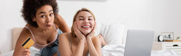 African american woman with credit card grimacing near happy friend and laptop in bedroom, banner — Stock Photo