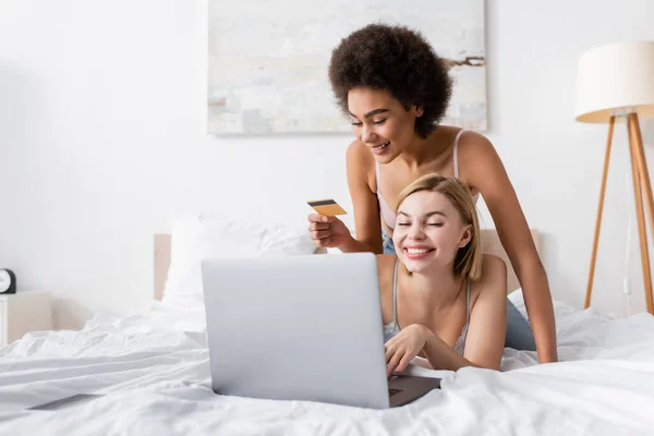 Heureuse femme afro-américaine tenant carte de crédit près ami souriant en utilisant un ordinateur portable sur le lit — Photo de stock