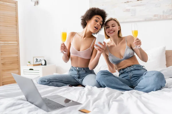 Amazed and cheerful multiethnic women holding champagne glasses with cocktails near blurred laptop on bed — Stock Photo