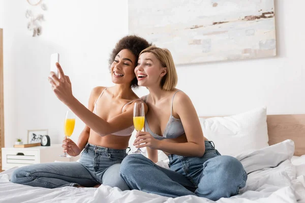Young and happy interracial women with cocktails in champagne glasses sitting on bed and taking selfie on cellphone — Stock Photo