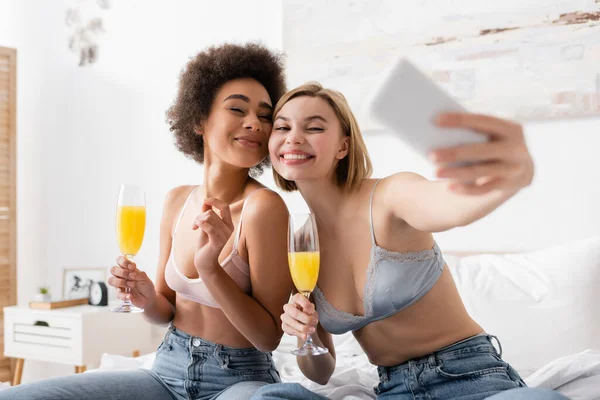Happy interracial women in lingerie and jeans holding champagne glasses with cocktails and taking selfie on blurred smartphone — Stock Photo