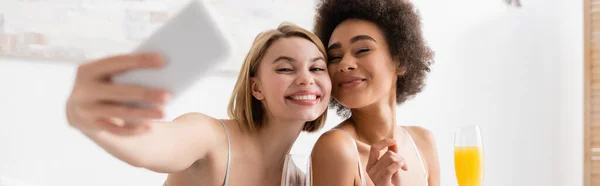 Happy blonde woman taking selfie with african american friend holding champagne glass with orange juice, banner — Stock Photo