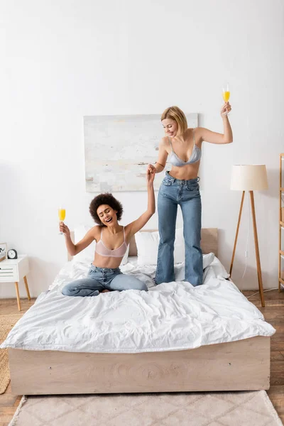 Happy blonde woman holding hands with african american friend sitting on bed with cocktail in champagne glass — Stock Photo