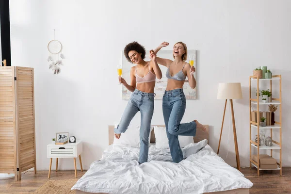 Full length of joyful interracial women in bras and jeans cocktails and dancing on bed — Stock Photo