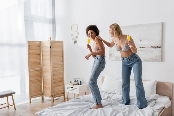 Full length of young multiethnic women in jeans and bras holding cocktails while standing on bed — Stock Photo