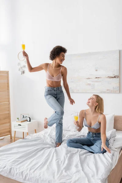 Joyful african american woman in bra and jeans posing with cocktail near blonde friend sitting on bed — Stock Photo