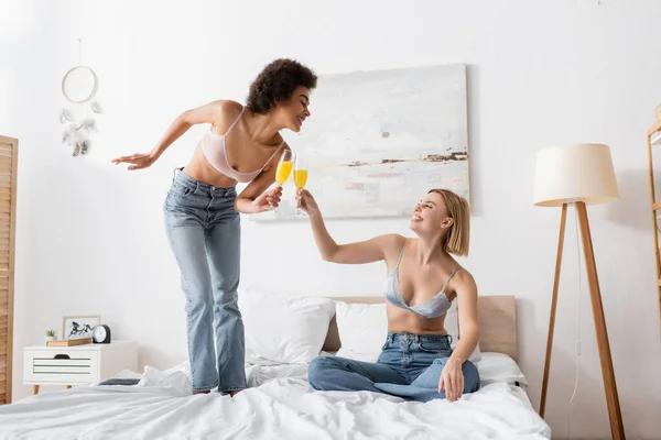 Joyful interracial women in jeans and bras clinking champagne glasses with cocktails in bedroom — Stock Photo