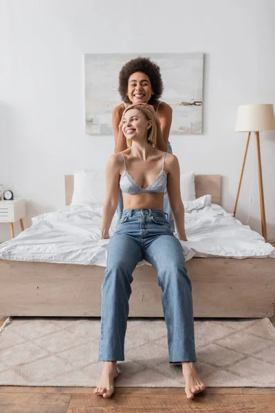 Happy african american woman with closed eyes touching head of blonde friend in bra and jeans sitting on bed and looking away — Foto stock