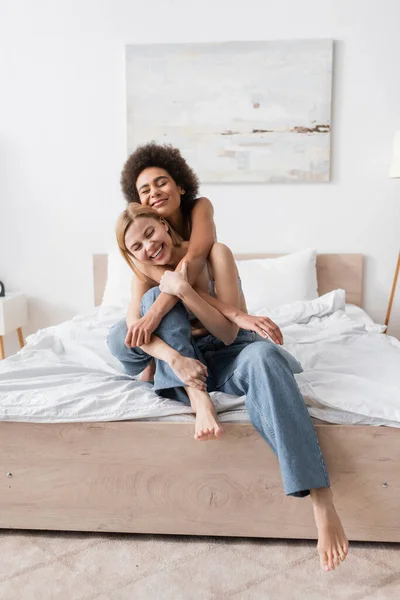 Happy african american woman embracing blonde barefoot friend in jeans sitting on bed at home — Stock Photo