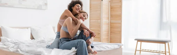 Joyful african american woman embracing blonde friend sitting on bed in bra and jeans, banner — Stock Photo