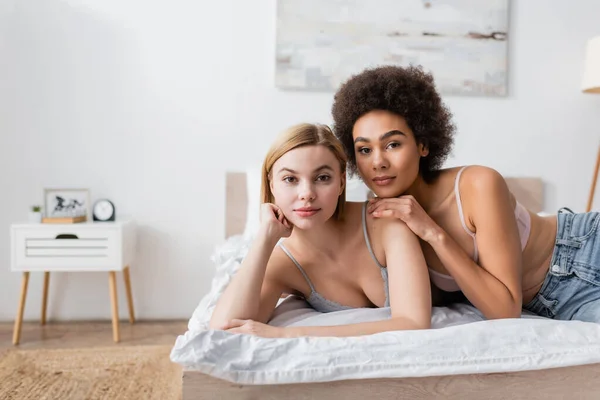 Young and pretty interracial women looking at camera while lying on bed at home — Stock Photo