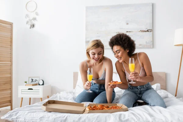 Happy interracial women holding champagne glasses with cocktails while looking at delicious pizza on bed at home — Stock Photo