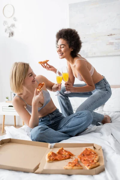 Joyful multiethnic friends in bra and jeans clinking champagne glasses with cocktails near tasty pizza on bed — Stock Photo