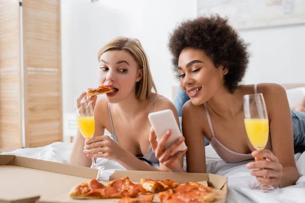 Donna afro-americana sorridente che tiene lo smartphone vicino a un amico biondo che mangia deliziosa pizza in camera da letto — Foto stock