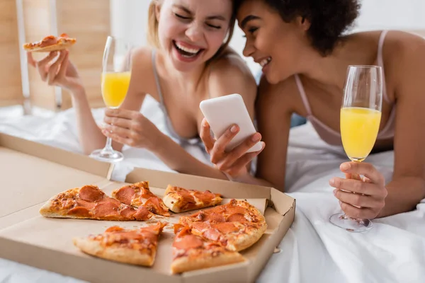 Excited multiethnic women with cocktails laughing near smartphone and tasty pizza in bedroom — Stock Photo